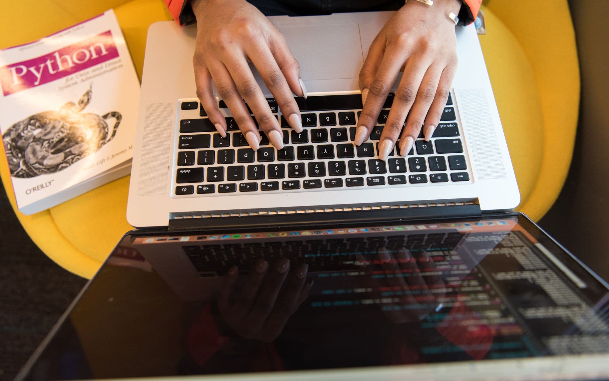 Close-up of hands typing code on a laptop beside a Python book
